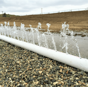Perforated pipe spouting multiple streams of water across a gravel bed in a groundwater recharge basin. The steep dirt side of the basin, topped with a chain link fence, is in the background.