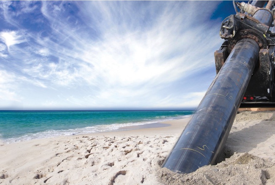 A sandy beach close to the shore line with a drilling rig in the foreground installing a slant well pipe. The drill rig pipe, which is approximately 12" in diameter, is buried in the sand up to a chalk mark reading 5'.