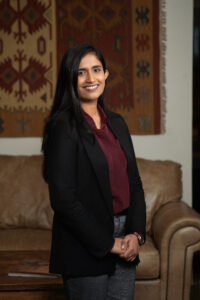 Bidisha is wearing a dark burgundy shirt under a black blazer and gray pants. She has long, dark hair and is standing with her hands clasped in front of her in a room with a leather cough behind her and a dark, patterned rug hanging on a light cream wall. 